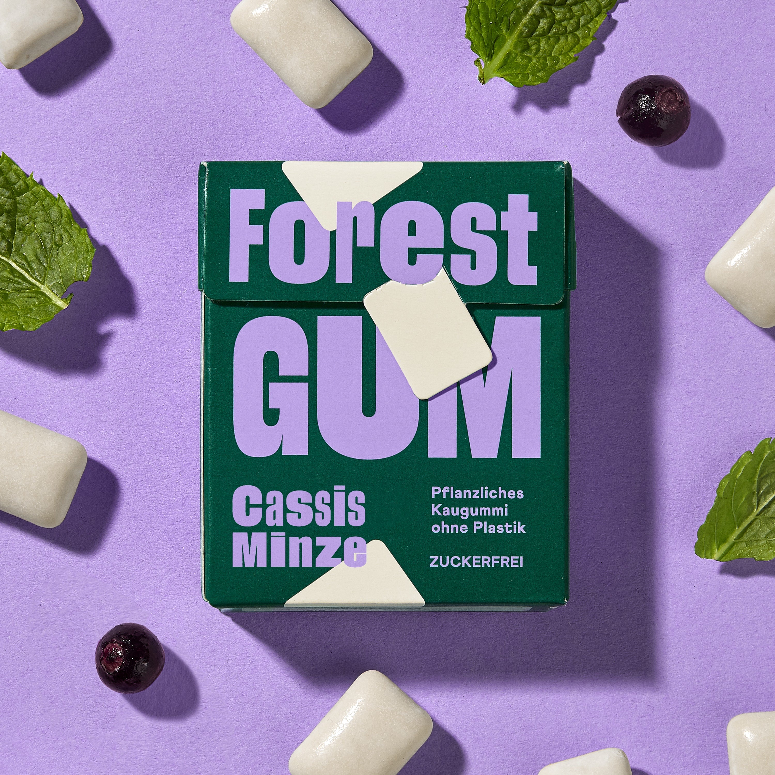 Forest Gum Cassis Minze Kaugummi-Packung, umgeben von Kaugummis, Johannisbeeren und Minzblätter.
